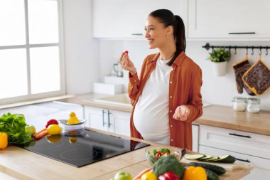 Mutfağın içinde sağlıklı yemek salatası hazırlarken taze sebze yiyen aşırı neşeli hamile kadın.