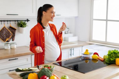Mutfağın içinde sağlıklı yemek salatası hazırlarken taze sebze yiyen aşırı neşeli hamile kadın.