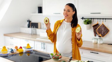 Hamilelik sırasındaki vitaminler. Mutlu hamile kadın avokado gösterirken sağlıklı sebze salatası hazırlıyor, tezgahta duruyor ve kameraya gülümsüyor.
