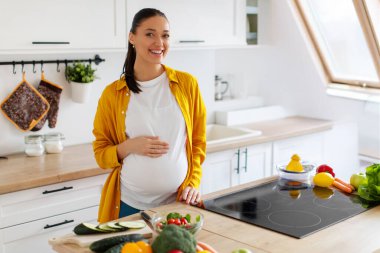 Sağlıklı beslenme ve annelik. Mutlu hamile kadın koca göbeğine dokunuyor ve kameraya gülümsüyor. Yemek hazırlarken mutfakta duruyor.