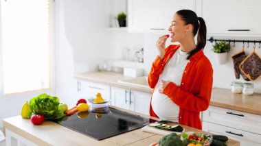 Mutfağın içinde salata pişirirken organik sebze yiyen genç ve tatmin olmuş bir kadın. Sağlık hizmeti, çocuk beklentisi, lezzet dilekleri.