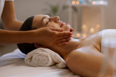 Kaplıca salonundaki sakin atmosferde rahatlatıcı yüz ve kafa masajı alan kadın, masör kadın çenesine ve yanaklarına dokunuyor, yakın plan.