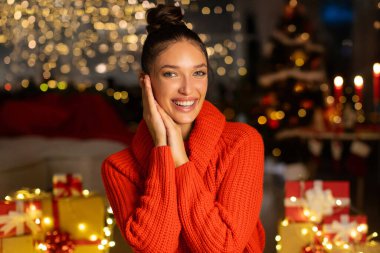 Kırmızı kazaklı mutlu Avrupalı bayan Noel süveteri içinde oturuyor ve kameraya gülümsüyor, çelenk ışıkları, mumlar ve arka planda bulanık Xmas ağacı