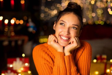 Mutlu hayalperest kadının portresi Noel ağacının ve çelenk ışıklarının önünde oturuyor, yana bakıyor ve gülümsüyor.