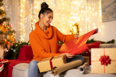 Heyecanlı bayan paketleme ve paketleme hediye kutuları, el işi kağıtları, dekore edilmiş odada kanepe ve arka planda Noel ağacı ile oturmak