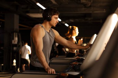 Genç kaslı adam spor salonundaki koşu programını seçiyor, kulaklık takıyor, antrenmana hazır.