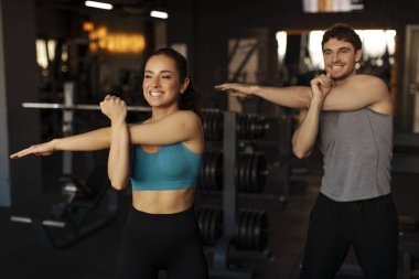 Mutlu adam ve kadın ısınıyor ve spor salonunda egzersiz yapmadan önce kollarını esnetiyorlar. Spor giysi giyiyorlar ve gülümsüyorlar.