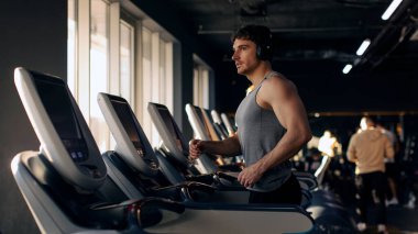 Kulaklık takan genç adam spor salonunda spor aletleriyle çalışıyor. Koşu bandında, panoramada, boş alanda koşuyor.