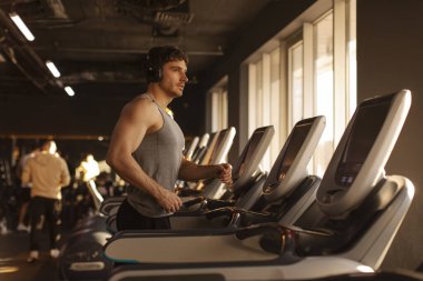 Kulaklık takan genç bir adam spor salonunda spor aletleriyle çalışıyor koşu bandında koşuyor, yan görüş, fotokopi çekiyor.
