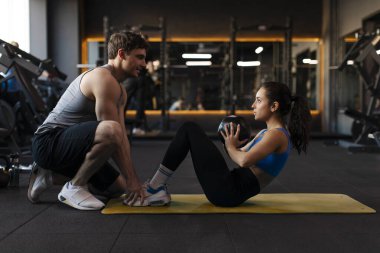 Erkek antrenör spor salonunda genç bayana yardımcı oluyor, kadın top tutarken mekik çalışıyor, tekniğine odaklanıyor, yan görüş atışı yapıyor.