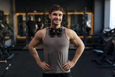 Spor giyim ve kulaklık giymiş güçlü kaslı bir adamın portresi modern spor salonunda duruyor ve kameraya gülümsüyor.