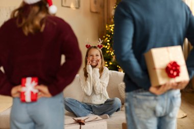 Aileler arkalarında Noel hediyesi kutuları saklıyorlar. Kızlarına sürpriz hazırlıyorlar. Kanepede oturuyorlar.