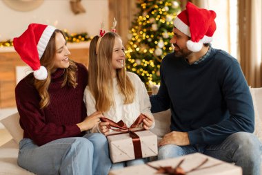 Noel Baba şapkalı heyecanlı ebeveynler genç kızlarına Noel 'i evde kutlayan ailelerine Noel hediyesi veriyor, kanepede oturuyor ve konuşuyorlar.