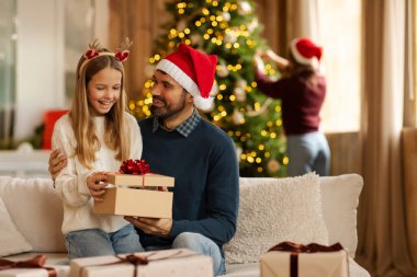 Kış bayramı kutlaması. Mutlu baba-kız Noel hediyesini açıyor, koltukta oturuyor ve anne arka planda Noel ağacını süslüyor.