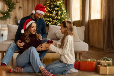 Şaşırmış anneye hediye kutusu veren, Noel arifesinde hediyelerini veren, Noel ağacıyla süslenmiş olarak oturma odasında oturan bir kız.