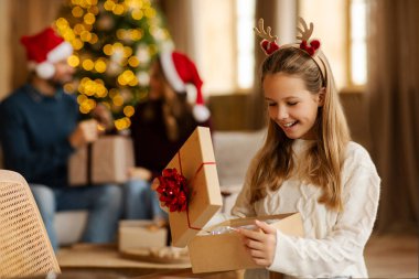 Noel için dekore edilmiş rahat oturma odasında hediyesini açarken geyik boynuzu takan heyecanlı kız. Ebeveynler arka planda koltukta oturup konuşuyorlar.