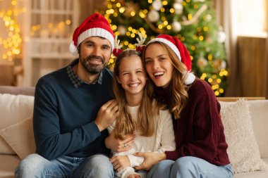 Üç kişilik Avrupalı aile neşeli bir şekilde gülümserken, şenlikli tatil şapkaları takıyor, güzel ışıklandırılmış Noel ağacıyla süslenmiş rahat oturma odasında kanepede oturuyor.