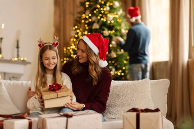 Mutlu anne-kız hediyelerini açarken baba da oturma odasında Noel ağacını şenlikli süslemelerle süslüyor.