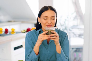 Tatmin olmuş bir kadın cam bardaktan taze sıkılmış smoothie kokan, masada yeşil meyve ve sebzelerle oturan,