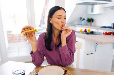 Zevkle pizza yiyen ve zevk alan mutlu bir kadın, parmaklarını kapalı gözlerle yalayan, mutfak masasında oturan, bedava alan.