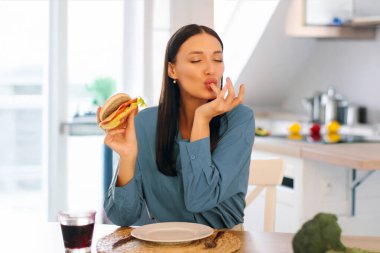 Lezzetli hamburger yerken parmağını yalayan, dışarıdan yemek sipariş eden, evde lezzetli hamburgerlerin tadını çıkaran genç bir bayan.