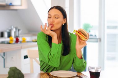 Lezzetli hamburger yerken parmağını yalayan, dışarıdan yemek sipariş eden, evde lezzetli hamburgerlerin tadını çıkaran genç bir bayan.