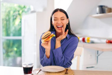 Heyecanlı genç bir kadın lezzetli hamburgerden ve dudaklarını silmekten zevk alıyor, evde abur cubur yiyor, mutfağın içinde oturuyor,