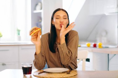 Tatmin olmuş, lezzetli hamburger yiyen, öğle yemeğinde hile yapan, hazır yemek sipariş eden ve sağlıklı olmayan atıştırmalıkların tadını çıkaran bir kadın.