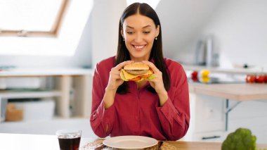 Aç kadın elinde kocaman bir hamburger, gülümsüyor, fast food yemekten ve evde hile yapmaktan zevk alıyor, mutfakta iç kısımda oturuyor.