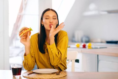 Tatmin olmuş, lezzetli hamburger yiyen, öğle yemeğinde hile yapan, hazır yemek sipariş eden ve sağlıklı olmayan atıştırmalıkların tadını çıkaran bir kadın.