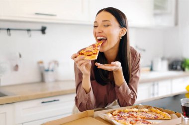 Pizzanın keyfi. Lezzetli pizzadan hoşlanan heyecanlı bir kadın, kapalı gözlerle pizza yerken, mutfakta masada otururken. Hile yemek, teslimat hizmeti konsepti.