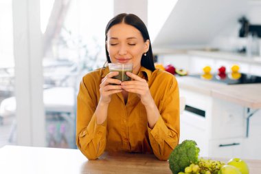 Tatmin olmuş bir kadın cam bardaktan taze sıkılmış smoothie kokan, masada yeşil meyve ve sebzelerle oturan,