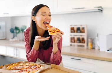 Pizzanın keyfi. Lezzetli pizzadan hoşlanan heyecanlı bir kadın, kapalı gözlerle pizza yerken, mutfakta masada otururken. Hile yemek, teslimat hizmeti konsepti.