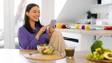 Pozitif genç bayan taze salata yiyor ve cep telefonu kullanıyor. Sağlıklı bir akşam yemeği yerken mutfakta oturuyor.