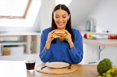 Aç kadın elinde kocaman bir hamburger, gülümsüyor, fast food yemekten ve evde hile yapmaktan zevk alıyor, mutfakta iç kısımda oturuyor.