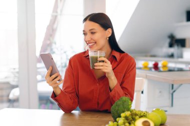 Mutlu bayan taze yeşil smoothie içiyor ve akıllı telefona bakıyor, mutfak masasında oturuyor, fotokopi çekiyor. Sağlıklı yemek, uygulama günlüğü