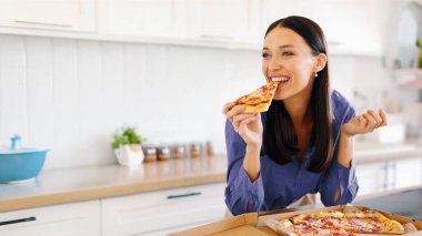 Mutlu kadın bir dilim lezzetli pizzayı ısırıyor, evde hile yemeğinin tadını çıkarıyor, mutfakta masada oturuyor. Pizza servisi konsepti