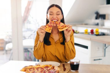 Heyecanlı bayan ev yapımı arka pizzayı seviyor, iki lezzetli dilimi ısırıyor, mutfak masasında oturuyor, fotokopi çekiyor. Sağlıksız beslenme aldatma yemeği