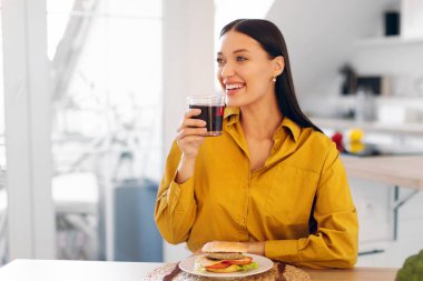 Evde lezzetli hamburgerler yerken meyve suyu içen tatmin olmuş bir kadın, mutfakta masada oturuyor, hazır yemek servisinin keyfini çıkarıyor, fotokopi çekiyor.