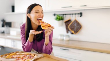 Pizzanın keyfi. Lezzetli pizzadan hoşlanan heyecanlı bir kadın, kapalı gözlerle pizza yerken, mutfakta masada otururken. Hile yemek, teslimat hizmeti konsepti.
