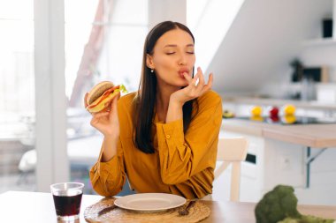 Lezzetli hamburger yerken parmağını yalayan, dışarıdan yemek sipariş eden, evde lezzetli hamburgerlerin tadını çıkaran genç bir bayan.