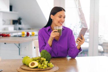 Mutlu bayan taze yeşil smoothie içiyor ve akıllı telefona bakıyor, mutfak masasında oturuyor, fotokopi çekiyor. Sağlıklı yemek, uygulama günlüğü