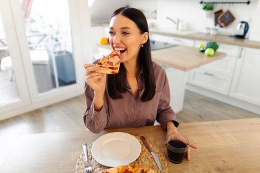 Mutlu bir kadının pizza yemesi, mutfakta masada oturması, evde aldatma yemeğinin tadını çıkarması. Abur cubur konsepti