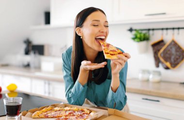 Pizzanın keyfi. Lezzetli pizzadan hoşlanan heyecanlı bir kadın, kapalı gözlerle pizza yerken, mutfakta masada otururken. Hile yemek, teslimat hizmeti konsepti.
