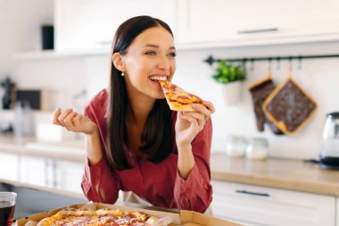 Mutlu kadın bir dilim lezzetli pizzayı ısırıyor, evde hile yemeğinin tadını çıkarıyor, mutfakta masada oturuyor. Pizza servisi konsepti