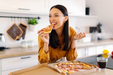 Mutlu kadın bir dilim lezzetli pizzayı ısırıyor, evde hile yemeğinin tadını çıkarıyor, mutfakta masada oturuyor. Pizza servisi konsepti