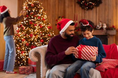 Afrikalı Amerikalı baba ve oğlu kanepede oturuyor, yılbaşı hediyesini birlikte açıyorlar, arka planda bayramlık Noel ağacı süsleyen kadın.
