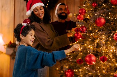 Heyecanlı Latin aile babaları ve oğlu Noel ağacını birlikte süslemenin keyfini çıkarıyorlar. Oda şenlikli ışıklar ve kırmızı süslemelerle dolu.