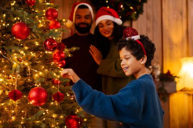 Afro-Amerikan ailesi Noel ağacını parlak ışık ve süslemelerle süslüyor, etrafı Noel süsleriyle çevrili.