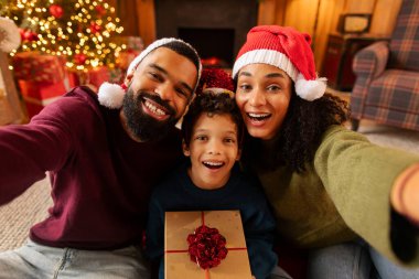 Neşeli Latin ailesi Noel arifesinde sıcak dekore edilmiş oturma odasında birlikte poz veriyor, selfie çekiyor ve mutlu bir şekilde gülümsüyor, Noel Baba şapkaları takıyor, paketlenmiş bir çocuk